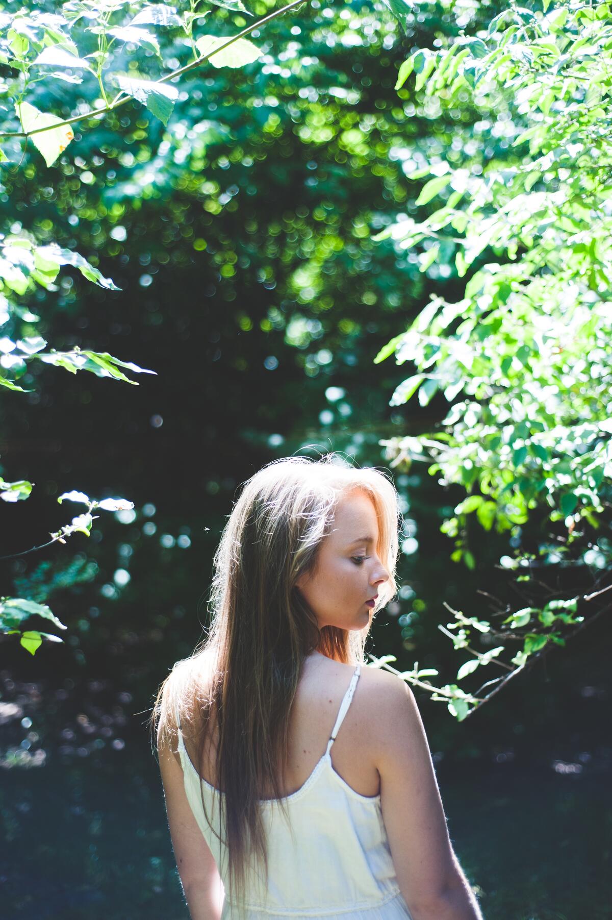 Светловолосая девушка стоит спиной на фоне деревьев