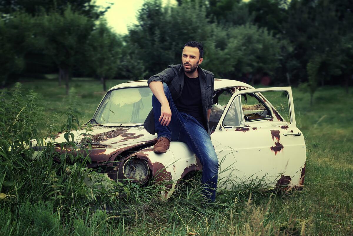 A guy with a beard sits on an abandoned car
