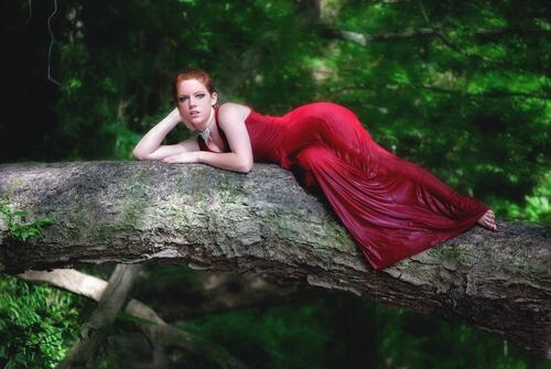 A red-haired girl in a red dress lying on a tree trunk