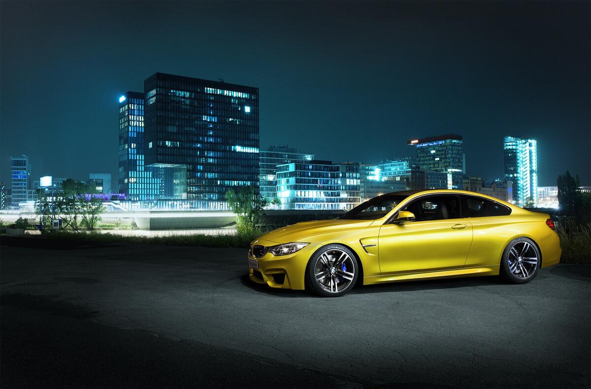 A gold BMW F82 M4 stands against the backdrop of a city at night